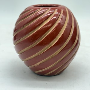 May include: A small, round, terracotta-colored vase with a spiral design. The vase has a glossy finish and is accented with lighter, tan-colored grooves that wrap around the body. The opening is circular.