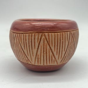 Puede incluir: Un pequeño cuenco de cerámica hecho a mano con un esmalte marrón rojizo y un patrón geométrico tallado en la superficie.