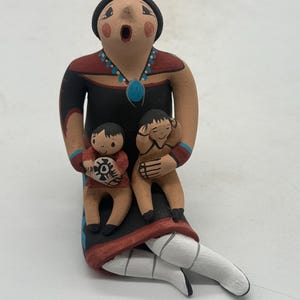 May include: A handcrafted ceramic figurine depicting a woman with two children. The woman wears a black dress with red and blue accents, a beaded necklace, and a black hat. The children are holding objects. The figurine is painted in earthy tones.