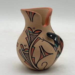 May include: A tan-colored ceramic pitcher with a curved spout and a rounded body. The pitcher is decorated with black, red, and turquoise geometric designs and a stylized snake. The rim and spout are a reddish-orange color.