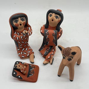 Puede incluir: Cuatro figuras de terracota. Dos figuras visten vestidos estampados, una sostiene un objeto rectangular. Un bebé descansa sobre una manta estampada y un pequeño ciervo está cerca. Las figuras tienen cabello negro y rasgos faciales.