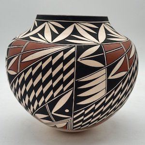 Può includere: Un vaso in ceramica fatto a mano di forma arrotondata. Il vaso presenta un bordo nero ed è decorato con intricati motivi geometrici in nero, crema e marrone rossiccio. Il design include forme a foglia e linee diagonali.
