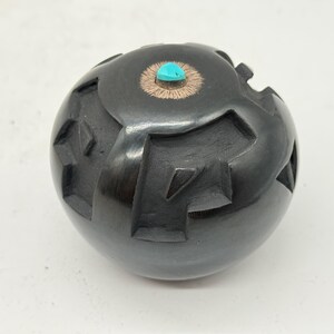 May include: A black, spherical pottery vessel with geometric cutouts. The top features a turquoise stone set in a copper-colored circle. The vessel has a glossy finish and a handcrafted appearance.