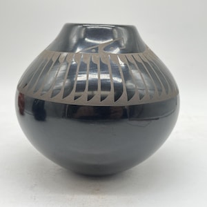 May include: A black ceramic vase with a rounded body and a narrow neck. The vase features a decorative band of geometric patterns in a lighter color around the upper portion. The vase has a glossy finish and sits on a white surface.