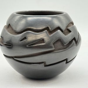 May include: A dark grey ceramic vase with a rounded form. The vase has a carved, geometric pattern around its exterior. The design consists of zigzags and angular shapes, creating a textured surface. The vase has a smooth, polished finish, and is likely handmade.