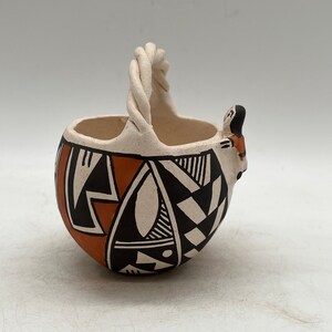 Puede incluir: Una pequeña cesta de cerámica blanca con un asa tejida y un diseño geométrico en negro, naranja y blanco. La cesta tiene una pequeña figura estilizada en el lateral.