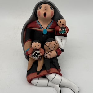 Puede incluir: Figurilla de cerámica pintada a mano de una mujer con tres hijos. La mujer viste un vestido negro con detalles rojos y turquesas, un tocado gris y mallas blancas. Los niños sostienen objetos con diseños geométricos.