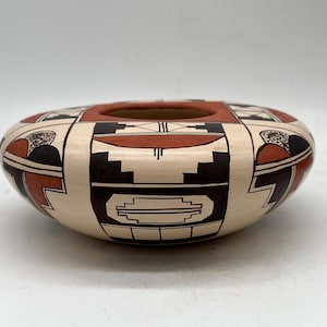 May include: A hand-painted, oval-shaped pottery bowl with a central opening. The bowl features a cream-colored base with geometric designs in black, red, and brown. The design includes stylized figures and patterns, showcasing traditional craftsmanship.