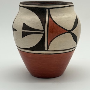 May include: A hand-painted ceramic vessel with a rounded body and a wide opening. The lower portion is a solid terracotta red, while the upper section features a cream-colored background adorned with geometric black and terracotta red patterns.