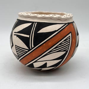 Puede incluir: Un pequeño cuenco redondo de cerámica blanca con un diseño geométrico negro y naranja. El cuenco tiene un borde ligeramente elevado.