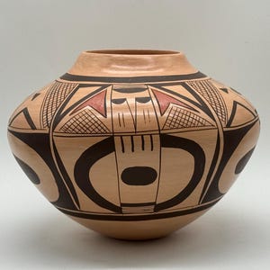 May include: A rounded ceramic vessel with a wide body and a narrow opening. The surface is a light tan clay colour, decorated with bold black geometric patterns, including circles, lines, and cross-hatching, accented by small terracotta red triangles.