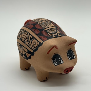 Pode incluir: Uma estatueta de porco em cerâmica bege com acabamento fosco, decorada com padrões geométricos pretos e vermelhos intrincados nas costas. O porco tem olhos pretos pintados à mão com cílios longos, sobrancelhas vermelhas e um pequeno focinho vermelho.