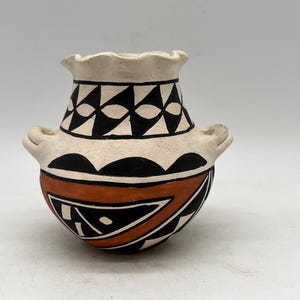 Puede incluir: Una pequeña olla de cerámica blanca con diseños geométricos negros y naranjas. La olla tiene dos pequeñas asas a los lados.