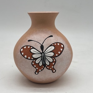 May include: A small, terracotta-colored vase with a white butterfly design. The butterfly has brown wings with white spots and a black body.