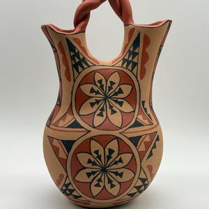 May include: A terracotta-colored clay vessel featuring a twisted handle connecting two spouts. The surface is decorated with hand-painted geometric patterns in black and dark red, including two prominent circular floral motifs on the front.