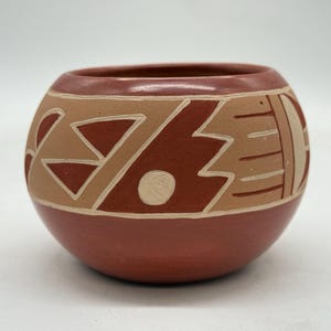May include: A round, hand-painted ceramic pot with a reddish-brown base and a lighter tan upper section. The pot features geometric designs in white and dark red, including triangles, zigzags, and curved lines. The rim is a darker shade of brown.