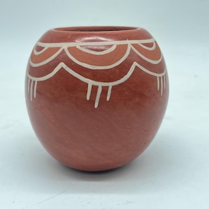 以下が含まれることがあります： テラコッタ色の小さな丸いセラミック花瓶。花瓶は、上部に白いとオレンジ色の曲線が描かれた装飾的な模様が特徴です。デザインは、様式化された波またはホタテの模様を連想させます。花瓶は滑らかでマットな仕上がりです。