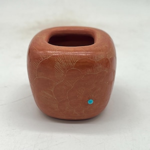 May include: A terracotta-colored, square-shaped vase with a carved floral design. The vase has a turquoise accent and a square opening at the top. The carved design covers the majority of the vase's surface.