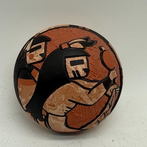 Pode incluir: Uma tigela de cerâmica redonda, cor de terracota, com um desenho preto e bege. A tigela apresenta figuras estilizadas com cabelo e roupas pretas. As figuras estão sobre um fundo de terracota. A tigela tem uma forma lisa e arredondada.