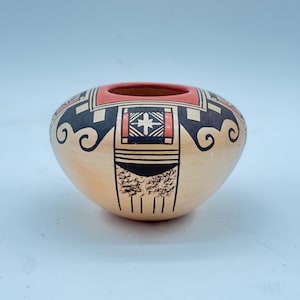 May include: A small, rounded ceramic bowl with a cream-colored base featuring intricate hand-painted geometric patterns in black and red. The design includes stylized scrollwork, rectangular motifs with cross-hatch details, and textured black accents.
