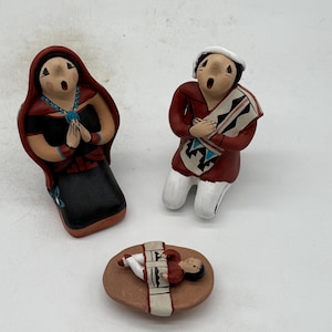 以下が含まれることがあります： 手描きのセラミック製降誕シーンの置物。赤いショールを着た女性、赤いシャツを着た男性、茶色の台座に横たわる赤ちゃんが含まれています。置物には詳細な顔の特徴と伝統的な衣装が施されています。