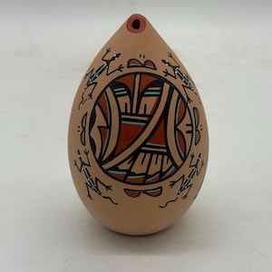 May include: A tan-colored, teardrop-shaped ceramic flute with a central geometric design in red, black, and turquoise. The flute is adorned with black frog figures around the central design. The top has a small hole for blowing.