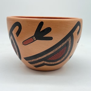 May include: A terracotta-colored ceramic bowl with black and red geometric designs. The bowl features stylized bird or feather motifs. The interior and exterior are the same color. The bowl has a rounded shape and a wide opening.