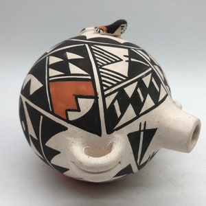 Puede incluir: Una olla de cerámica blanca con diseños geométricos negros y naranja. La olla tiene un pico y una pequeña figura de pájaro en la parte superior.