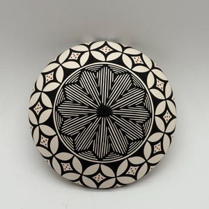 May include: A round ceramic plate featuring a central black and white geometric star pattern surrounded by a border of interlocking circles. Each circle contains a small brown diamond shape with four dots, set against a stark white background.