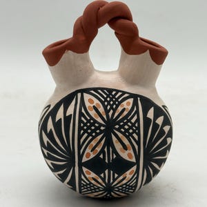 Könnte beinhalten: Handgefertigte Keramikvase mit cremefarbenem Korpus und einem dekorativen geometrischen Muster in Schwarz und Orange. Die Vase hat einen gedrehten Griff in Rotbraun und zwei Öffnungen oben.