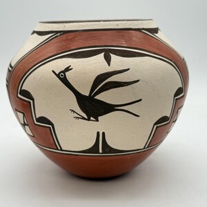 Può includere: Vaso in ceramica dipinto a mano con un disegno di roadrunner. Il vaso presenta uno sfondo color crema con un'illustrazione di roadrunner nera. Il bordo e la base sono marrone-rossastro, con motivi geometrici in bianco e nero.