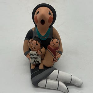 May include: A hand-painted ceramic figurine depicting a woman holding two children. The woman wears a black outfit with a turquoise necklace and a black head covering. One child holds a sign that reads "Pueblo Stories". The figurine is painted in earth tones.