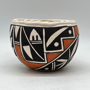 Puede incluir: Un pequeño cuenco de cerámica pintado a mano con un diseño geométrico en negro, blanco y naranja. El cuenco tiene una forma ligeramente redondeada y un borde blanco.