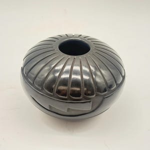 May include: A polished black ceramic vessel with a rounded, low-profile shape. The top section features a series of vertical, carved grooves radiating from a central circular opening, while the lower body displays a smooth, matte geometric pattern.