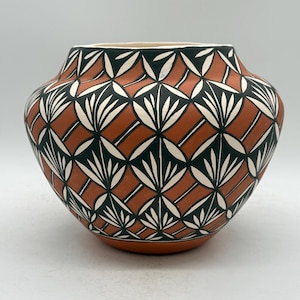 May include: A terracotta-coloured ceramic vase with a geometric pattern. The design features black and white leaf-like shapes set against a terracotta background. The vase has a wide body and a slightly flared rim.