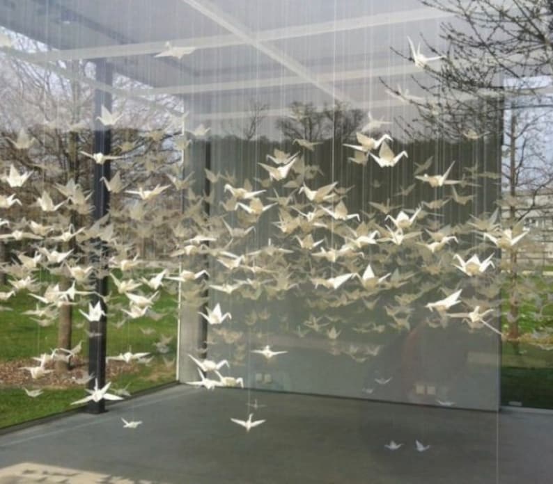 May include: A large room with a glass ceiling and walls, filled with hundreds of white origami paper cranes hanging from the ceiling.