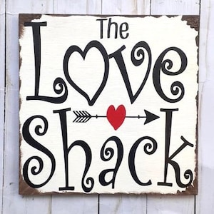 Puede incluir: Un letrero de madera blanco con letras negras que dice "The Love Shack" con un corazón rojo y una flecha en el medio.