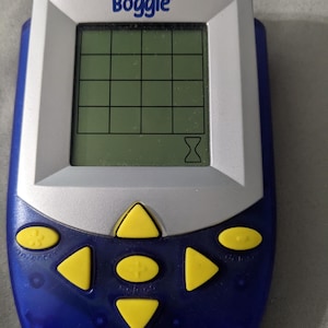 May include: A handheld Boggle game with a blue and silver casing. The screen displays a grid for letter placement, with the word "Boggle" printed at the top. Yellow directional buttons and other function buttons are located below the screen.