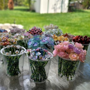 Puede incluir: Una colección de coloridos ramos de flores de cuentas en vasos de plástico transparente. Las flores son de varios colores, incluyendo rojo, rosa, morado y azul. Los tallos son verdes.