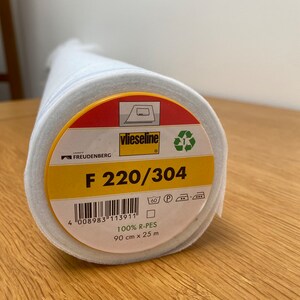 Puede incluir: Un rollo de tela blanca con una etiqueta amarilla y roja. La etiqueta dice "vlieseline" y "F 220/304". También tiene un código de barras e instrucciones de lavado y planchado.