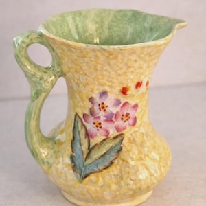 May include: A small, yellow ceramic jug with a green handle and interior. The jug is decorated with pink and purple flowers and green leaves. The handle and interior have a mottled green pattern.