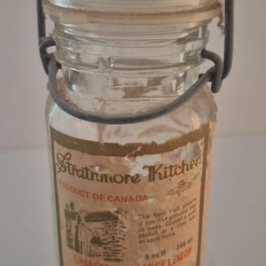 Puede incluir: Tarro de vidrio vintage con asa y tapa de alambre metálico. La etiqueta dice "Strathmore Kitchen" y "Mermelada de Pomelo Naranja Limón". El tarro contiene 256 ml de mermelada y es un producto de Canadá.