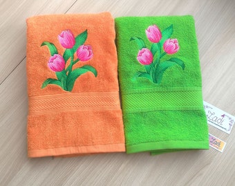 May include: Two embroidered towels with pink flowers on a light wood surface. The towels are folded and one is orange and the other is green.