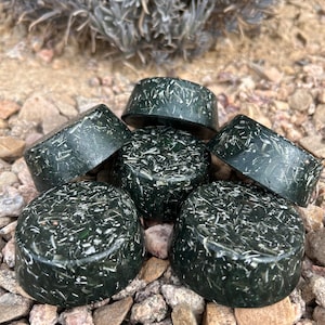 May include: Five dark green round soap bars with a speckled texture. The soap bars are arranged in a pile on a bed of small brown rocks.
