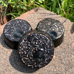 May include: Three round black soap bars with embedded silver and copper flakes. The soap bars have a swirl design in the center.