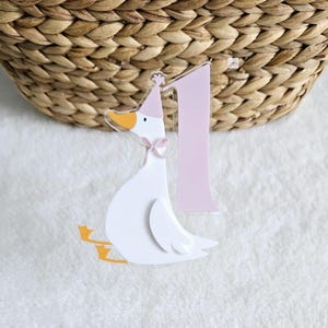 May include: A white goose wearing a pink party hat and holding a pink number one. The goose is holding the number one in front of it with its wings spread out.
