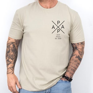 Puede incluir: Una camiseta beige con un dise&ntilde;o gr&aacute;fico negro que presenta las letras "PAPA" en un c&iacute;rculo con una cruz. El texto "Jack Maya Est. 2020" est&aacute; debajo del gr&aacute;fico.