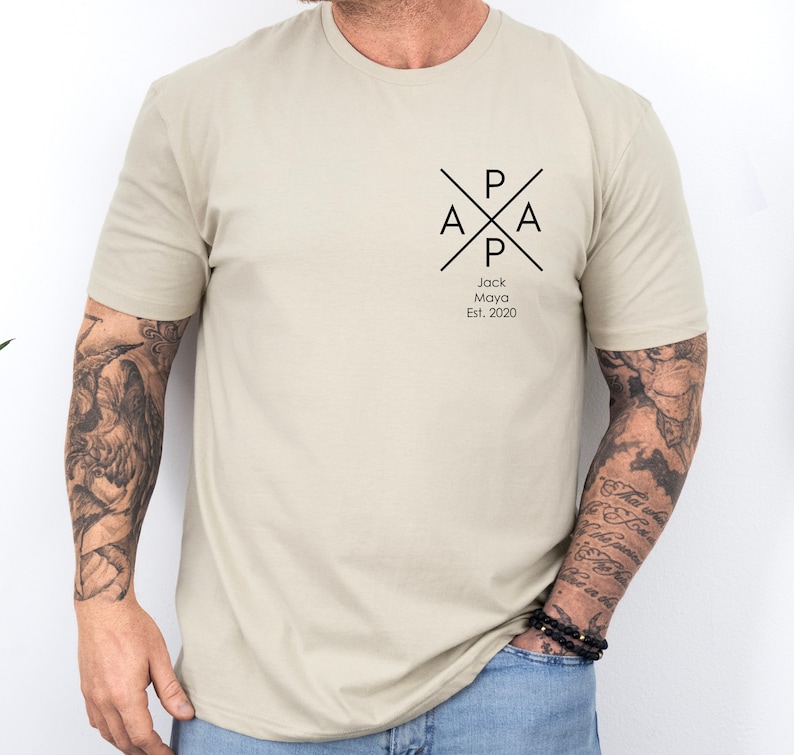 Puede incluir: Una camiseta beige con un dise&ntilde;o gr&aacute;fico negro que presenta las letras "PAPA" en un c&iacute;rculo con una cruz. El texto "Jack Maya Est. 2020" est&aacute; debajo del gr&aacute;fico.