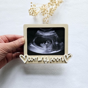 May include: A wooden frame holding a sonogram image. The frame features a heart design and the words "coming soon" in white script. The image is held by a hand.