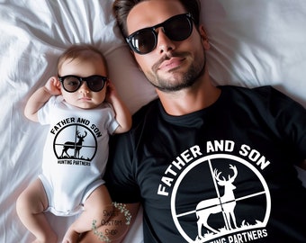 May include: A white onesie and a black t-shirt with a deer in a target design. The text on the onesie reads "Father and Son Hunting Partners". The text on the t-shirt reads "Father and Son Hunting Partners".